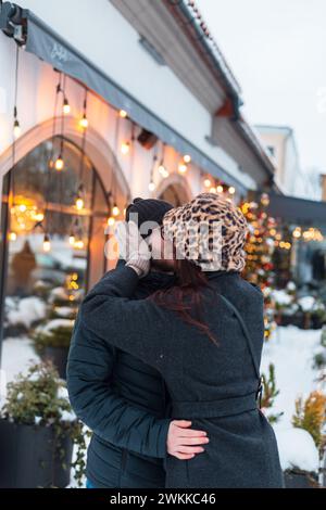 Mode schöne junge Liebende in trendigen Winterkleidern Stände und küssen in der Stadt in der Nähe eines Gebäudes mit Lichtern. Mädchen küsst den Kerl Stockfoto