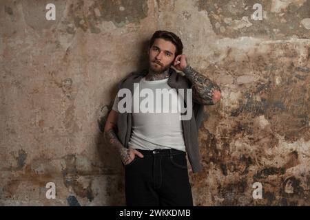 Ein cooles, stylisches, junges, hübsches Hipster-Modell mit Tattoos in modischer Kleidung, Strickpullover und weißem T-Shirt steht neben einer alten Grunge-Wand Stockfoto