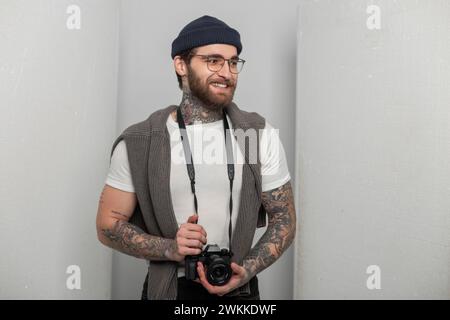 Glücklicher gutaussehender professioneller Fotograf Mann mit Lächeln mit Tattoo mit Strickmütze und Pullover, der spiegellose Fotokamera hält und in Studi steht Stockfoto