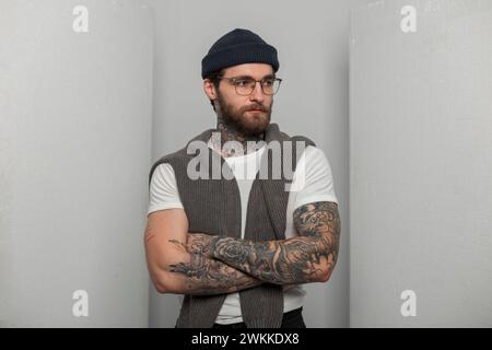 Cooler, gutaussehender Hipster-Mann mit Bart, Schnurrbart und Tattoo mit stylischer Brille, Strickmütze in einem modischen weißen T-Shirt und einem Pullover im Th Stockfoto