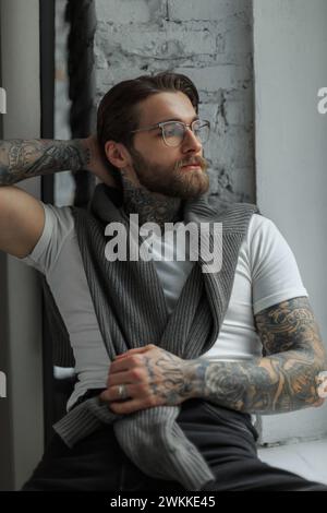 Der coole Hipster-gutaussehende Kerl mit Bart und Frisur mit Tattoo und Brille in einem weißen T-Shirt mit Pullover sitzt und entspannt sich zu Hause in der Nähe des Sieges Stockfoto