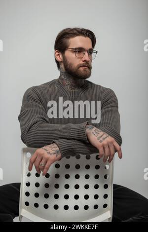 Cooler, hübscher junger Hipster-Mann mit Frisur und Bart mit Brille in einem modischen Strickpullover sitzt auf einem weißen Stuhl im Studio Stockfoto