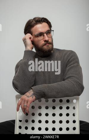 Cooles Hipster-hübsches Mann-Model mit Frisur und Bart mit Brille in einem modischen Strickpullover sitzt auf einem weißen Stuhl im Studio Stockfoto