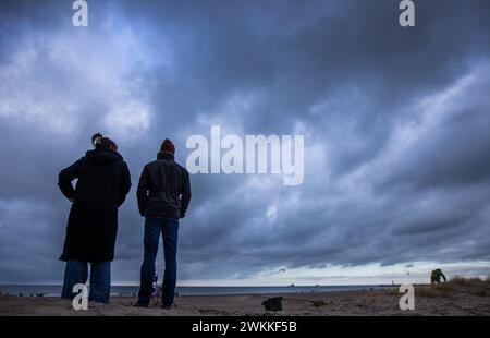 Rostock, Deutschland. Februar 2024. Wanderer stehen an einem Strandzugang in Warnemünde vor einem Hintergrund dunkler Wolken. Meteorologen erwarten starke Winde mit Sturm an der Ostseeküste. Quelle: Jens Büttner/dpa/Alamy Live News Stockfoto