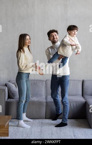 Ganzkörpervater und Mutter lächeln und heben gemischten Sohn nahe der Couch Stockfoto