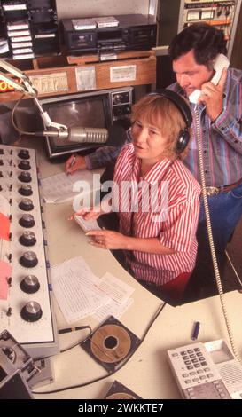 Copperas Cove Texas USA, 1996: Weibliche Disc Jockey spricht in Mikrofon, während ihr Mann und Co-Moderator während der Live-Übertragung im Kontrollraum eines Radiosenders in einer kleinen Stadt in Central Texas telefonieren. ©Bob Daemmrich Stockfoto