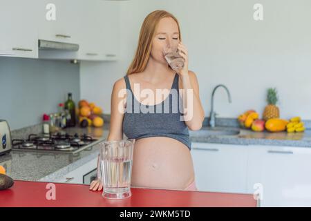 Eine schwangere Frau, die die lebenswichtigen Vorteile des Wassers während der Schwangerschaft berücksichtigt, steht mit einem Glas in der Küche und unterstreicht die entscheidende Rolle der Hydratation bei Stockfoto