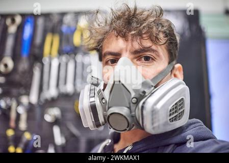 Porträt eines lateinischen jungen Arbeiters mit Atemschutzmaske und Blick in die Kamera in seiner Werkstatt. Echte Menschen bei der Arbeit. Stockfoto
