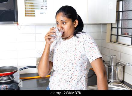indische Frau trinkt Wasser aus Glas in der Küche zu Hause und schaut in die Kamera Stockfoto