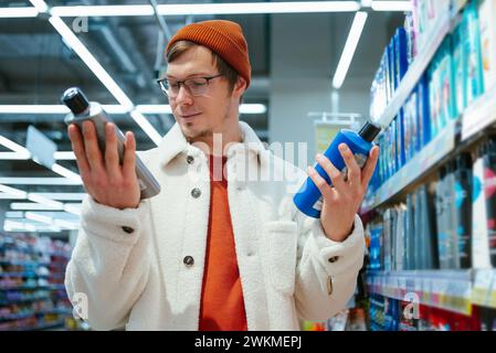 Ein anspruchsvoller Einkäufer in Fleecejacke und Mütze vergleicht zwei Shampoo-Flaschen in einem Ladengang, wobei seine Aufmerksamkeit von den Details auf den Etiketten erfasst wird. Stockfoto