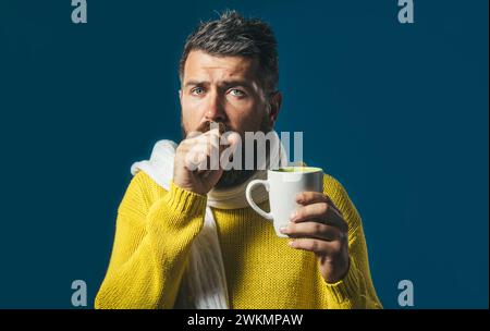 Mann hustet. Kranker Mann mit Erkältung oder Bronchitis, der in die Faust hustet. Bärtiger gutaussehender Mann in Pullover und Schal mit einer Tasse medizinischen Tee. Unwohlsein Stockfoto