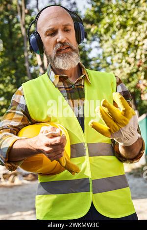 Fleißiger Hüttenbauer in Schutzhandschuhen und Weste posiert mit Kopfhörern und Helm vor Ort Stockfoto