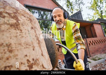 Gutaussehender, fröhlicher Mann in Sicherheitshandschuhen und Kopfhörern, der mit der Kreissäge arbeitet, Hüttenbauer Stockfoto