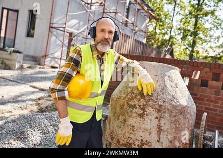 Fröhlicher Hüttenbauer in Schutzhandschuhen und Weste mit Kopfhörern und Helm, der in die Kamera lächelt Stockfoto