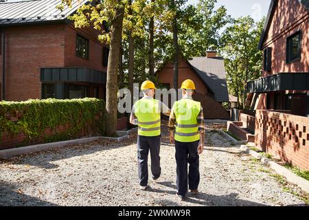 Rückansicht von engagierten Bauarbeitern in Sicherheitswesten mit Bauplan vor Ort, Hüttenbauer Stockfoto