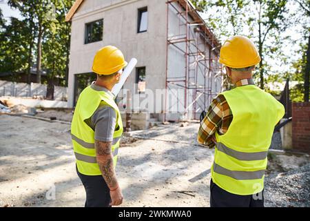 Rückansicht von hingebungsvollen Hüttenbauern in Schutzhelmen mit einem Entwurf, in dem ihre Arbeit vor Ort diskutiert wird Stockfoto