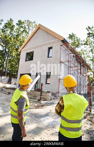 Rückansicht von hingebungsvollen Hüttenbauern in Schutzhelmen mit einem Entwurf, in dem ihre Arbeit vor Ort diskutiert wird Stockfoto