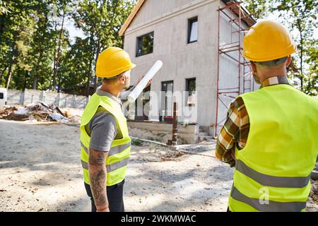 Rückansicht von hingebungsvollen Hüttenbauern in Schutzhelmen mit einem Entwurf, in dem ihre Arbeit vor Ort diskutiert wird Stockfoto