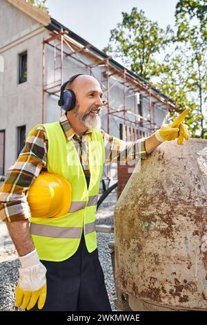 Fröhlicher Hüttenbauer in Schutzhandschuhen und Weste posiert mit Kopfhörern und Helm vor Ort Stockfoto