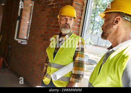 Gutaussehende Bauarbeiter in Schutzhelmen arbeiten vor Ort zusammen, Hüttenbauer Stockfoto