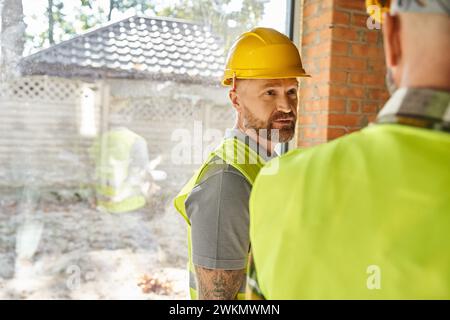 Konzentrieren Sie sich auf einen bärtigen Arbeiter im Schutzhelm, der mit seinem Kollegen auf der Baustelle, Bauherren, spricht Stockfoto