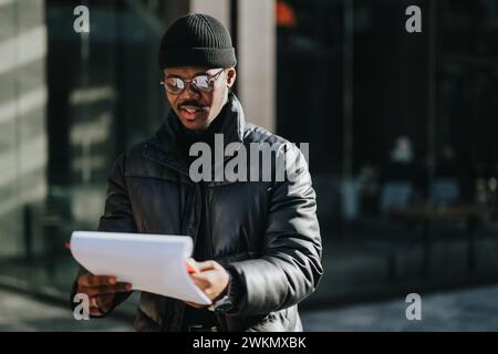 Ein junger, professioneller afroamerikanischer Mann in stilvoller Winterkleidung prüft sorgfältig Papierkram an einem sonnigen Tag in einer urbanen Umgebung. Stockfoto