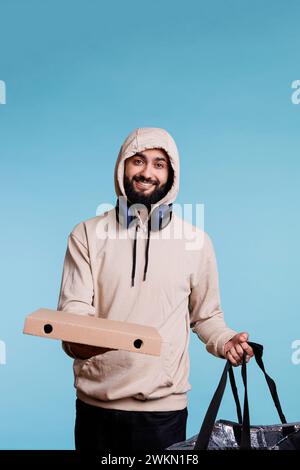 Lachender Essensdienst arabischer Kurier mit Pizzaschachtel und Rucksack Porträt. Fröhlicher junger Mann in der Kapuze, der Fastfood-Take-away-Bestellung gibt und mit sorglosem Ausdruck in die Kamera blickt Stockfoto