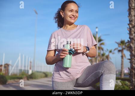Lächelnde, glückliche Athletin, entschlossene Sportlerin, die nach dem Sport am Strand Wasser trinkt. Stockfoto