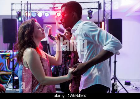 Ein romantisches junges Paar, das beim Clubben die Hände hält und zusammen tanzt. Vielfältige Freunde und Freundinnen feiern und ziehen auf der Tanzfläche in einem Nachtclub mit Lichtern Stockfoto