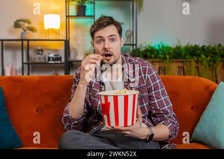 Glücklicher kaukasischer Mann isst Popcorn und schaut heimlich, feiert Erfolg, Siegesgewinn, Zielerreichung, sitzend auf Sofa im Wohnzimmer. Aufgeregt, junger Kerl, der Fäuste in die Kamera schaut Stockfoto