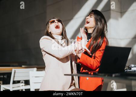 Zwei überfröhliche Geschäftsfrauen teilen sich einen unbeschwerten Moment mit Lachen, während sie an einem Laptop im Freien mit hellem Sonnenlicht arbeiten. Stockfoto