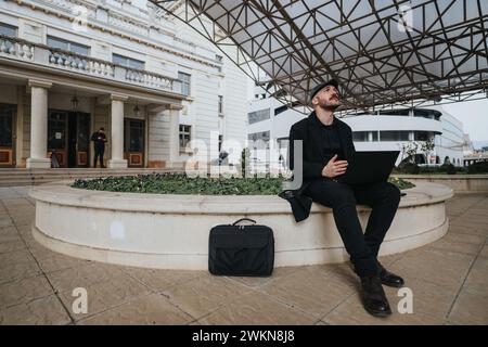 Geschäftsmann, der im Freien arbeitet und mit seinem Laptop in urbaner Umgebung nach oben blickt. Stockfoto