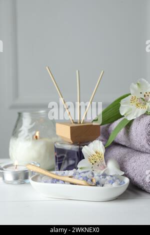 Spa-Zusammensetzung. Handtücher, brennende Kerzen, Schilflufterfrischer, Meersalz und schöne Blumen auf weißem Holztisch, Nahaufnahme Stockfoto