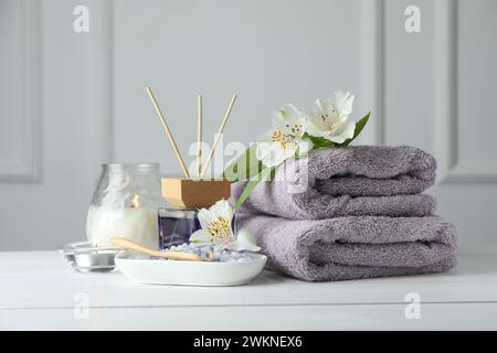 Spa-Zusammensetzung. Handtücher, brennende Kerzen, Schilflufterfrischer, Meersalz und wunderschöne Blumen auf weißem Holztisch Stockfoto