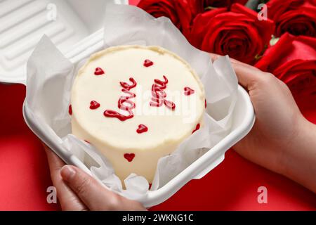 Frau, die eine Kiste mit bento-Kuchen am roten Tisch hält, Nahaufnahme. St. Valentinstag-Überraschung Stockfoto