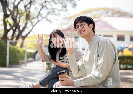 Zwei freundliche und fröhliche asiatische Freunde oder Kollegen winken mit den Händen, um ihre Freunde zu begrüßen und zu grüßen, während sie in einem Stadtpark chillen Stockfoto