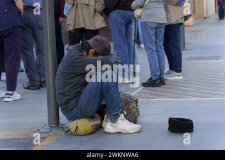 Obdachloser Mann auf der Straße bettelt um Almosen mit traurigem Ausdruck, Kopf zwischen den Beinen und eine Mütze auf dem Boden für Münzen Stockfoto