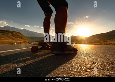 Die Silhouette aus nächster Nähe zeigt die Beine der Skater auf einem langen Brett auf der Bergstraße vor dem Sonnenuntergangshimmel Stockfoto
