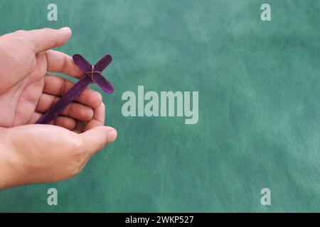 Männliche Hand, die lila hölzerne Kreuz hält, flach auf grünem Hintergrund. Heilige Woche und Feierlichkeiten in der Leihzeit Stockfoto