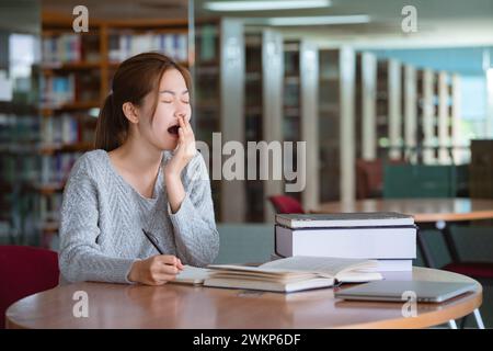 Junge asiatische Frau gähnt beim Lesen eines Buches in der Bibliothek. Stockfoto