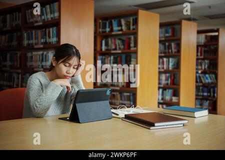 Gestresste junge Studentin sitzt am Holztisch und liest Buch während der Prüfungsvorbereitung in der Bibliothek. Stockfoto