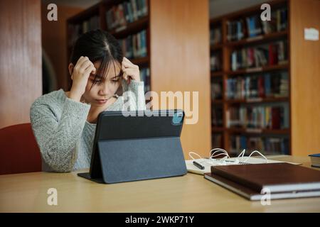 Gestresste junge Studentin sitzt am Holztisch und liest Buch während der Prüfungsvorbereitung in der Bibliothek. Stockfoto