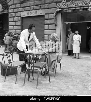 Vaction in Italien in den 1950er Jahren Eine junge Frau, die an einem Café-Tisch in einer geschäftigen Mailänder Straße sitzt. Ein Kellner steht neben ihr und ist höflich und verzaubert von der schwedischen Blondine. Mailand Italien 1950. Foto Kristoffersson Ref. DV19 *** Lokale Bildunterschrift *** © Classic Picture Library. Alle Rechte vorbehalten. Geschützt durch eine digitale Signatur. Bildüberwachung und -Schutz ist auf diesem Bild aktiviert. Die Lizenz, Präsentation oder Newsletter beinhaltet KEINE Nutzung in sozialen Medien. Stockfoto