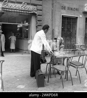 Vaction in Italien in den 1950er Jahren Eine junge Frau, die an einem Café-Tisch in einer geschäftigen Mailänder Straße sitzt. Ein Kellner steht neben ihr und ist höflich und verzaubert von der schwedischen Blondine. Mailand Italien 1950. Foto Kristoffersson Ref. DV19 *** Lokale Bildunterschrift *** © Classic Picture Library. Alle Rechte vorbehalten. Geschützt durch eine digitale Signatur. Bildüberwachung und -Schutz ist auf diesem Bild aktiviert. Die Lizenz, Präsentation oder Newsletter beinhaltet KEINE Nutzung in sozialen Medien. Stockfoto