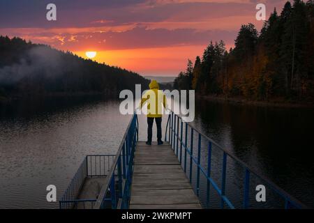 Mann, der auf der Brücke über dem See steht, in einem gelben Regenmantel. Blick auf Bozcaarmut bei Sonnenuntergang. Stockfoto