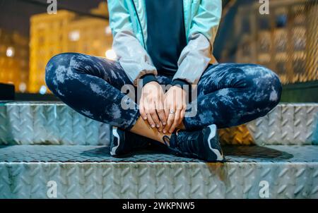 Nahaufnahme einer nicht erkennbaren Athletin, die nachts mit einem Kreuz auf einer Metalltreppe sitzt. Läuferin mit Laufleggings und Turnschuhen Stockfoto