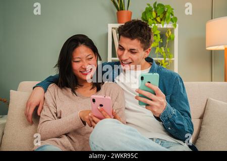 Echtes multirassisches Ehepaar, das ein Handy benutzt, um Apps auf dem Wohnzimmer-Sofa zu suchen. Junge Menschen, die lächeln und Spaß mit einem Smartphone haben. Zwei Freunde teilen sich in den sozialen Medien. Hochwertige Fotos Stockfoto