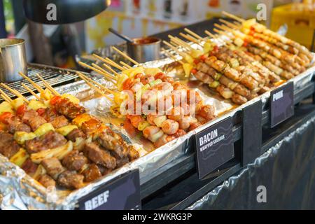 Eine appetitliche Auswahl an gegrillten Spießen mit Hühnchen, Schweinefleisch und mit Speck gewickelten Würstchen, begleitet von Gemüse, wird in einem Street Food stan präsentiert Stockfoto