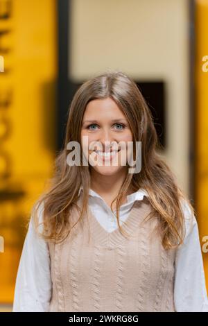 Eine junge Frau mit fließenden braunen Haaren lächelt in einer beigefarbenen Strickweste über einer weißen Bluse vor einem leuchtend gelben Hintergrund. Stockfoto