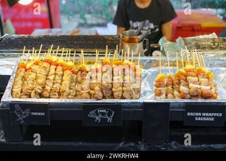 Eine appetitliche Auswahl an gegrillten Spießen mit Hühnchen, Schweinefleisch und mit Speck gewickelten Würstchen, begleitet von Gemüse, wird in einem Street Food stan präsentiert Stockfoto